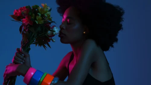 Foto de uma mulher negra de perfil. Ela está na frente de um fundo azul e está cheirando um buquê de flores. A mulher tem cabelo crespo e preto, usa várias pulseiras coloridas e está levemente iluminada por uma luz cor de rosa.