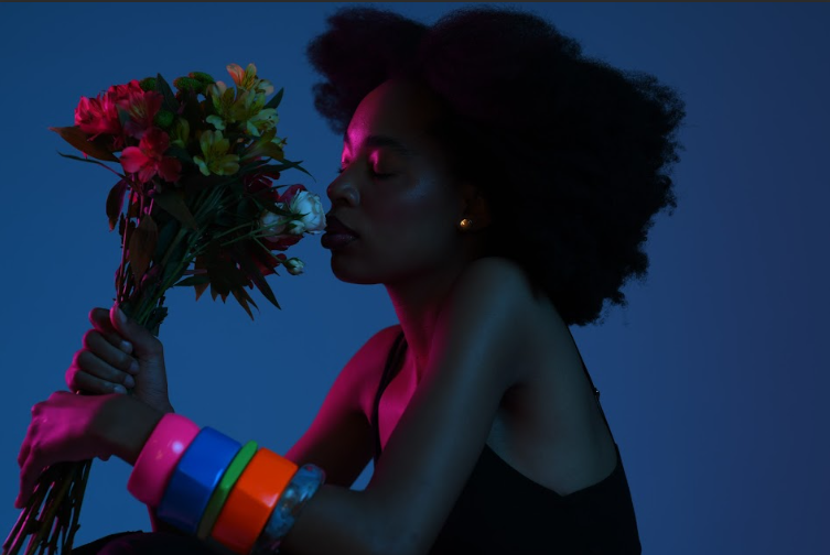 Foto de uma mulher negra de perfil. Ela está na frente de um fundo azul e está cheirando um buquê de flores. A mulher tem cabelo crespo e preto, usa várias pulseiras coloridas e está levemente iluminada por uma luz cor de rosa.