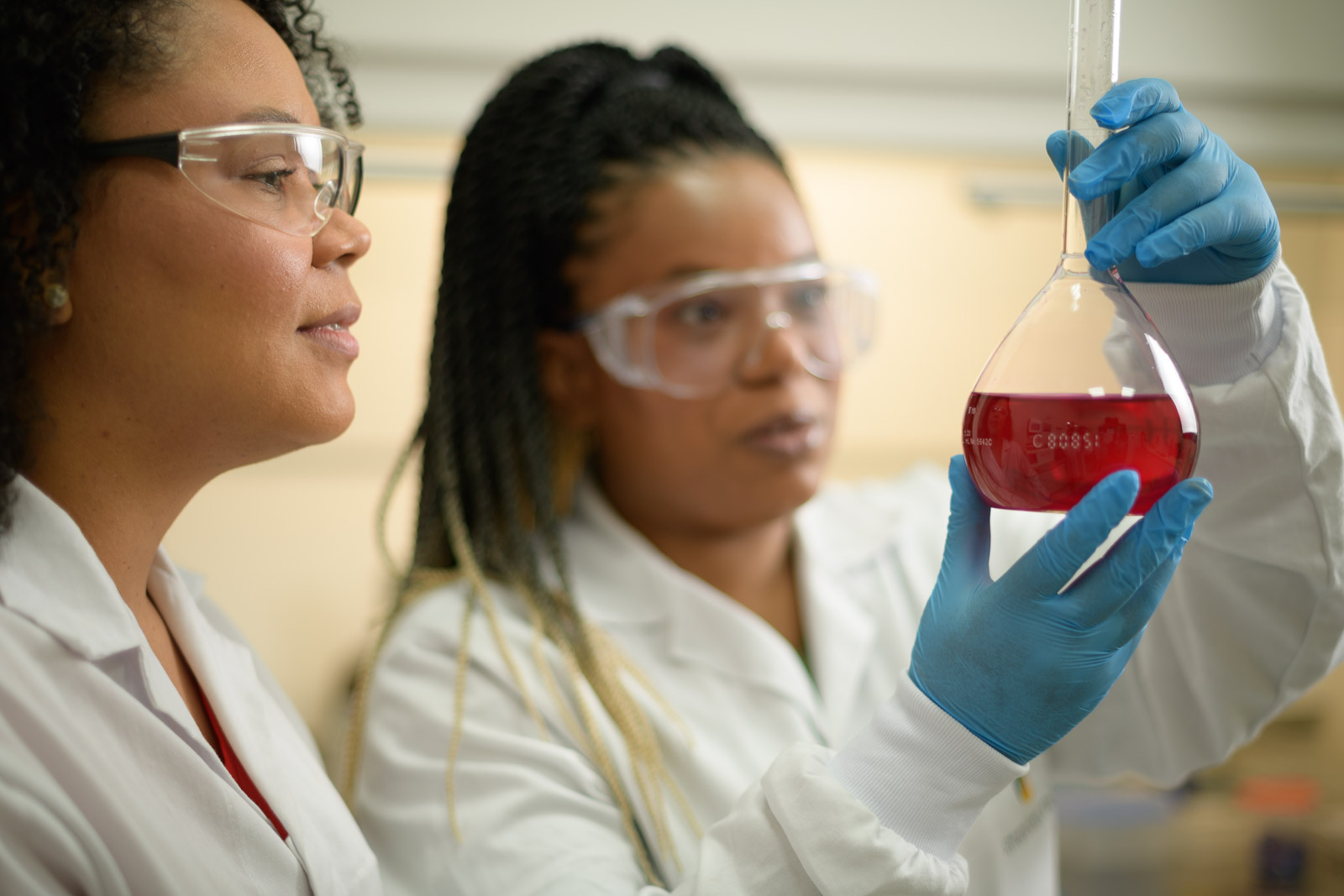 Duas mulheres negras e cientistas estão lado a lado em um laboratório. Elas estão usando óculos de proteção, jalecos branco e luvas azuis. A mulher da direita segura na altura do rosto um vidro de erlenmeyer com um líquido vermelho.