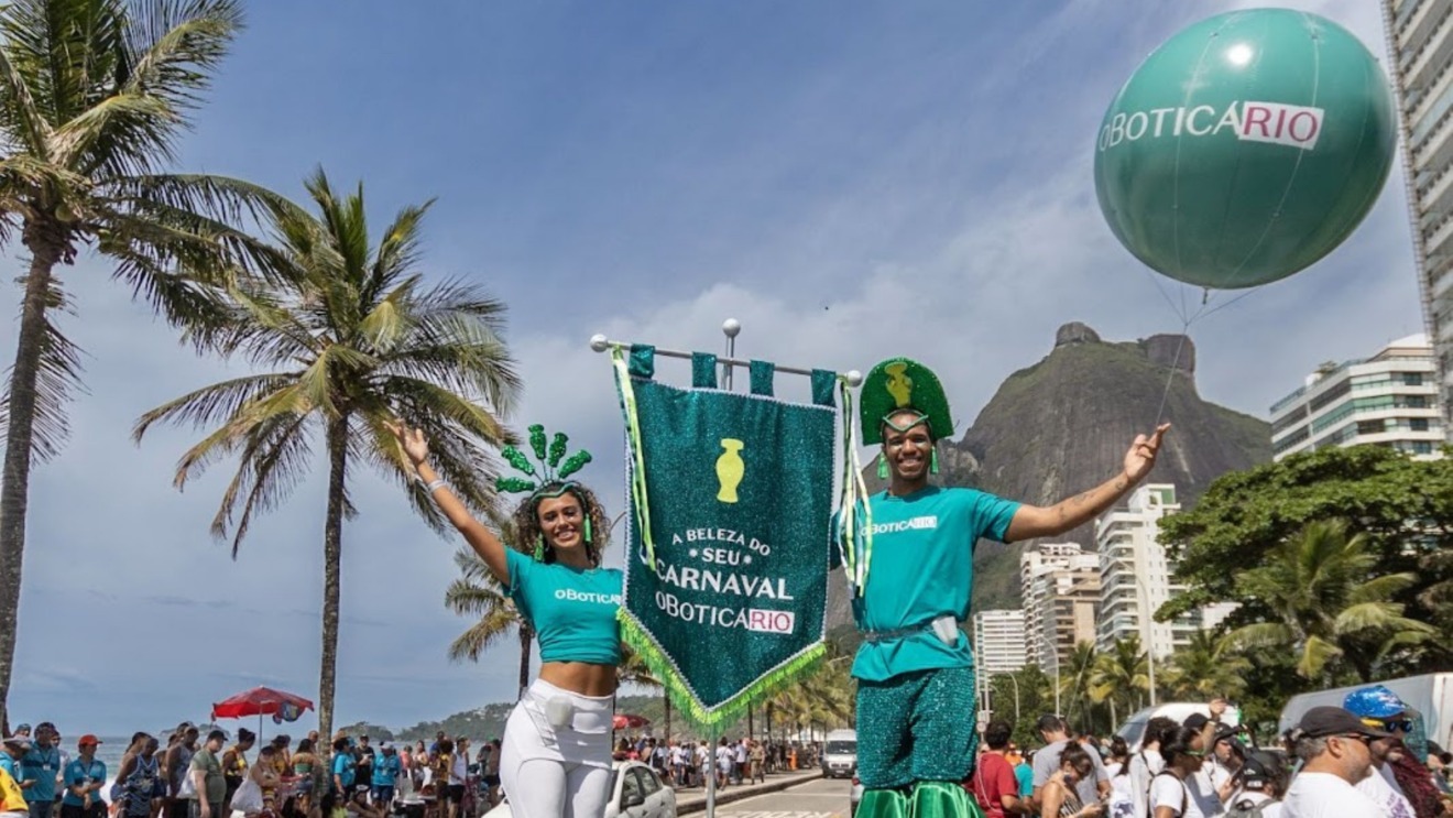 A imagem mostra uma das ações de Carnaval do Grupo Boticário. Na foto, um bloco está desfilando à beira mar no Rio de Janeiro, com destaque para dois foliões, um homem e uma mulher, que desfilam usam camisetas de cor verde com a marca de O Boticário e carregam um estandarte verde com o desenho de uma ânfora e a frase "A beleza do seu carnaval O Boticário".