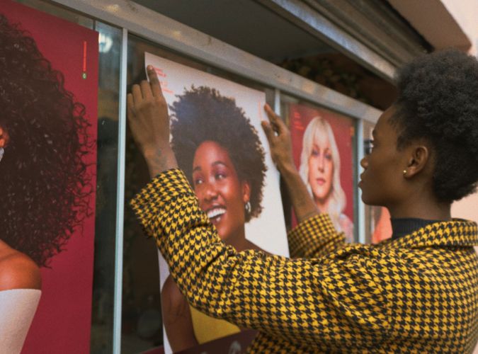 Imagem da nova campanha da Vult para nova linha de produtos para cabelos. Na imagem, uma mulher negra está colando o pôster, com a foto de uma mulher negra, no vidro à sua frente. 