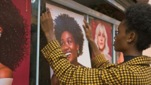 Imagem da nova campanha da Vult para nova linha de produtos para cabelos. Na imagem, uma mulher negra está colando o pôster, com a foto de uma mulher negra, no vidro à sua frente.