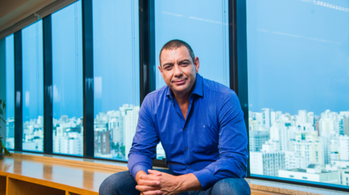 Sérgio Sampaio é o VP de Operações do Grupo Boticário. Ele é um homem negro, com olhos castanhos e cabelos raspados. Ele está usando uma camisa azul e calça jeans e está sentado em um móvel com uma janela ao fundo.