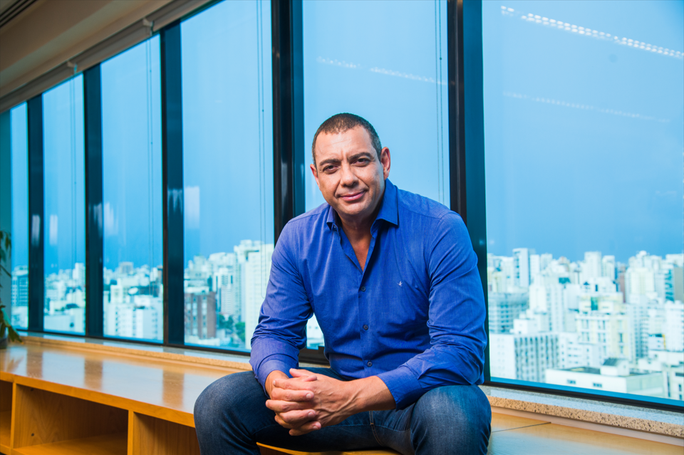 Sérgio Sampaio é o VP de Operações do Grupo Boticário. Ele é um homem negro, com olhos castanhos e cabelos raspados. Ele está usando uma camisa azul e calça jeans e está sentado em um móvel com uma janela ao fundo.