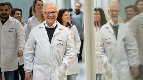 Imagem de Miguel Krigsner, fundador do Grupo Boticário, andando pelo laboratório de inovação da empresa. Miguel é um homem idoso, branco, de cabelos brancos e óculos de armação escura. Ele está sorrindo.