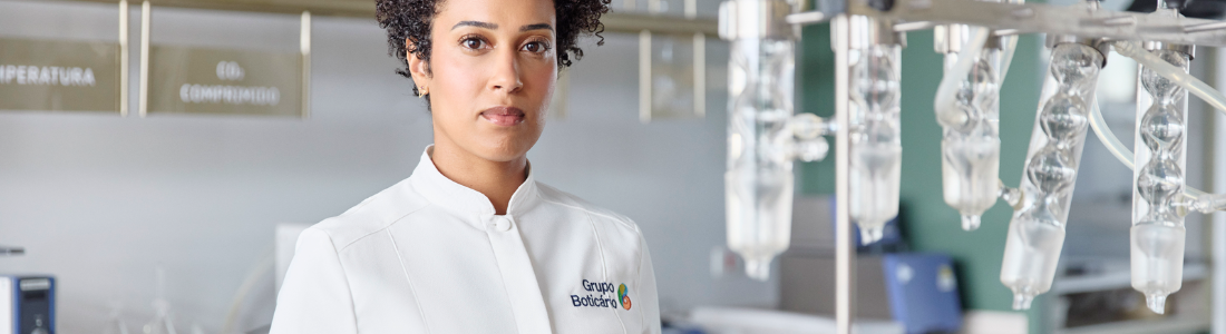 Uma mulher de pele morena e cabelos cacheados curtos, vestindo um jaleco branco com o logo do "Grupo Boticário", está em um laboratório. Ela olha diretamente para a câmera com uma expressão séria. Ao seu redor, há diversos equipamentos de laboratório de vidro, bancadas verdes e prateleiras com outros instrumentos científicos. A cena sugere um ambiente de pesquisa e desenvolvimento dentro da indústria de cosméticos ou produtos relacionados.