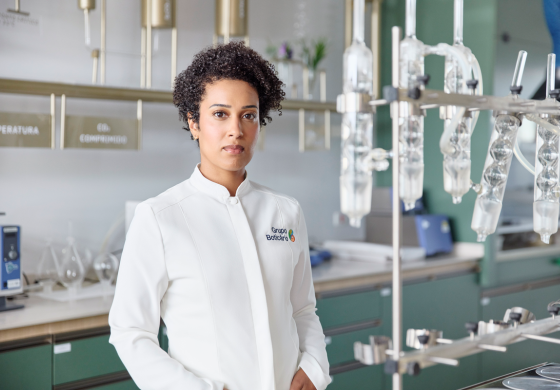 Uma mulher de pele morena e cabelos cacheados curtos, vestindo um jaleco branco com o logo do "Grupo Boticário", está em um laboratório. Ela olha diretamente para a câmera com uma expressão séria. Ao seu redor, há diversos equipamentos de laboratório de vidro, bancadas verdes e prateleiras com outros instrumentos científicos. A cena sugere um ambiente de pesquisa e desenvolvimento dentro da indústria de cosméticos ou produtos relacionados.