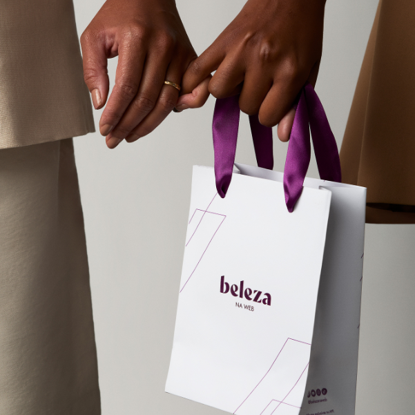 Duas mãos de tons de pele diferentes seguram as alças roxas de uma sacola de compras branca. A sacola exibe a inscrição 
