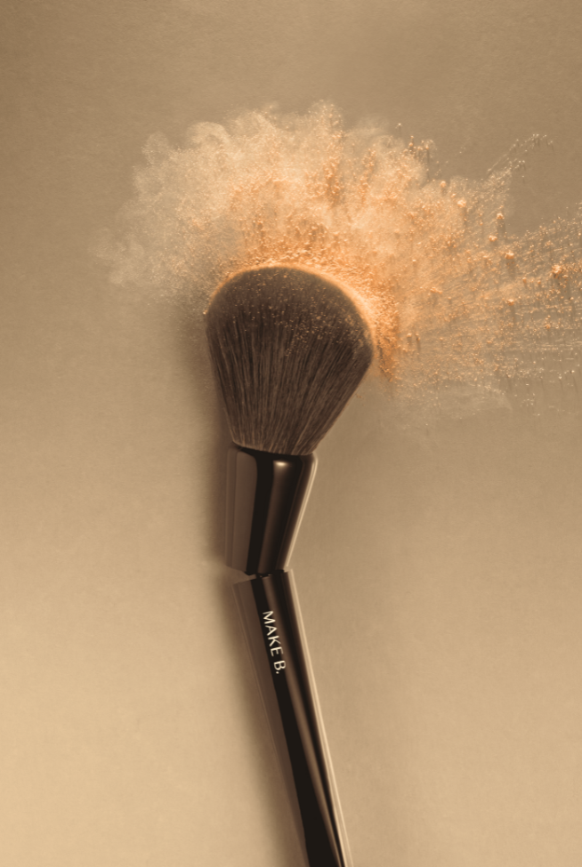 Um pincel de maquiagem preto da marca Make B. é capturado em close-up, com uma nuvem de pó solto bege explodindo de suas cerdas. O pincel está posicionado diagonalmente contra um fundo bege suave e uniforme. A luz incide sobre a cena, iluminando a textura macia das cerdas e a dispersão fina do pó no ar. O logotipo "MAKE B." é visível ao longo do cabo preto e brilhante do pincel. A imagem evoca a aplicação de maquiagem e a leveza dos produtos em pó.
