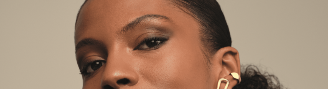 Um close-up do lado esquerdo do rosto de uma mulher de pele morena revela detalhes de sua maquiagem e acessórios. Seu olhar é direcionado ligeiramente para baixo, focando em seus olhos com sombra marrom esfumada e cílios alongados. Uma sobrancelha bem definida emoldura a pálpebra superior. Na orelha, ela usa dois brincos dourados distintos: um pequeno círculo na parte superior e um retângulo alongado pendurado no lóbulo. O fundo é um tom neutro e suave, concentrando a atenção nos traços faciais e nos adornos da mulher.