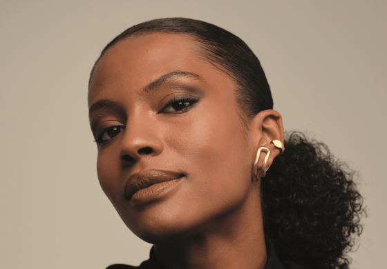 Um close-up do lado esquerdo do rosto de uma mulher de pele morena revela detalhes de sua maquiagem e acessórios. Seu olhar é direcionado ligeiramente para baixo, focando em seus olhos com sombra marrom esfumada e cílios alongados. Uma sobrancelha bem definida emoldura a pálpebra superior. Na orelha, ela usa dois brincos dourados distintos: um pequeno círculo na parte superior e um retângulo alongado pendurado no lóbulo. O fundo é um tom neutro e suave, concentrando a atenção nos traços faciais e nos adornos da mulher.