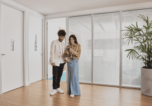Em um espaço de escritório moderno com piso de madeira clara, um homem e uma mulher estão lado a lado, olhando para um smartphone que a mulher segura. O homem veste um blazer branco sobre uma camiseta branca e calças pretas, enquanto a mulher usa uma jaqueta marrom clara e uma saia jeans longa e clara. Ao fundo, há divisórias de vidro com persianas brancas e um vaso grande com uma planta de folhas verdes. Portas brancas com discretas identificações de "Eudora" e "Grupo Boticário" são visíveis nas paredes laterais. A cena sugere uma interação digital casual em um ambiente de trabalho corporativo.