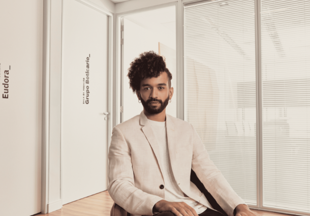 Imagem de um rapaz com blazer e camiseta bege sentado em cadeira no escritório do Grupo Boticário.