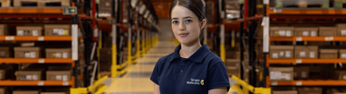 Uma jovem com cabelo escuro preso em um rabo de cavalo, vestindo uma camiseta polo azul escura com o logo do "Grupo Boticário", está no centro de um armazém. Ela olha diretamente para a câmera com um sorriso suave. Ao seu redor, há altas estantes metálicas laranjas e amarelas, repletas de caixas de papelão. O corredor é iluminado e sugere um ambiente de logística ou distribuição.