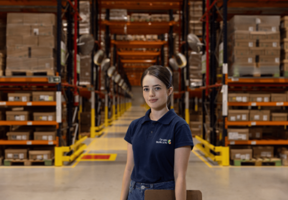Uma jovem com cabelo escuro preso em um rabo de cavalo, vestindo uma camiseta polo azul escura com o logo do "Grupo Boticário", está no centro de um armazém. Ela olha diretamente para a câmera com um sorriso suave. Ao seu redor, há altas estantes metálicas laranjas e amarelas, repletas de caixas de papelão. O corredor é iluminado e sugere um ambiente de logística ou distribuição.