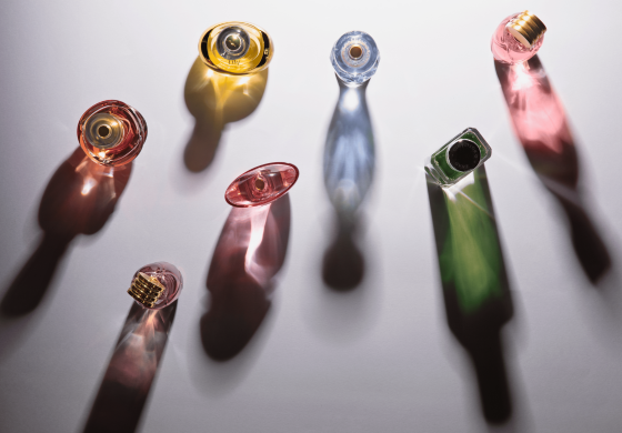 Frascos de perfume de diferentes formatos e cores são vistos de cima sobre uma superfície branca. Cada frasco projeta uma sombra alongada e colorida, criando um efeito visual de cáustica de refração da luz através do líquido e do vidro. Os frascos contêm líquidos em tons de dourado, azul claro, rosa, laranja, rosa vibrante e verde escuro. As tampas dos frascos variam em cor e design, adicionando um toque individual a cada um. A composição minimalista e a iluminação direta realçam as cores e as formas dos frascos e suas sombras.