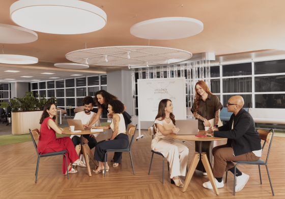 A imagem mostra um espaço de trabalho moderno e colaborativo com várias pessoas engajadas em diferentes atividades. O ambiente parece ser um escritório com iluminação suave proveniente de grandes luminárias circulares no teto. O chão é de madeira clara. Na parte esquerda da imagem, há um grupo de cinco pessoas reunidas em torno de uma mesa redonda. Quatro delas estão sentadas em cadeiras de design simples com estrutura metálica e assento de madeira clara, enquanto uma mulher está ligeiramente inclinada sobre a mesa, participando da conversa. Há laptops e cadernos sobre a mesa, sugerindo uma reunião de trabalho ou uma sessão de brainstorming. As pessoas neste grupo parecem estar interagindo ativamente, com expressões faciais que indicam engajamento e discussão. À direita, outra mesa redonda menor acomoda duas pessoas sentadas, cada uma com um laptop à sua frente. Eles também parecem estar trabalhando ou colaborando. Uma mulher de cabelos ruivos está de pé ao lado da mesa, parecendo estar envolvida na discussão ou apresentando algo. Ao fundo, há uma divisória de vidro com cortinas brancas translúcidas, separando esta área de outros espaços do escritório. É possível ver mais janelas grandes com persianas brancas e algumas plantas em vasos, indicando um ambiente de trabalho bem iluminado e com um toque de natureza. Uma lousa branca com algumas anotações rabiscadas é visível atrás da divisória de vidro. A atmosfera geral da imagem é de um ambiente de trabalho dinâmico, onde a colaboração e a comunicação são incentivadas. As pessoas parecem estar trabalhando em equipe de forma informal e flexível.