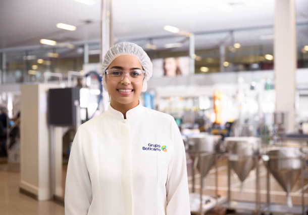 Uma jovem sorridente, vestindo um jaleco branco, touca e óculos de proteção, posa para a câmera em um ambiente industrial limpo e iluminado. O logo do "Grupo Boticário" está visível em seu jaleco. Ao fundo, equipamentos de produção em aço inoxidável e outras máquinas sugerem uma fábrica ou laboratório de cosméticos ou produtos farmacêuticos. A imagem transmite uma sensação de profissionalismo e trabalho em um ambiente controlado.