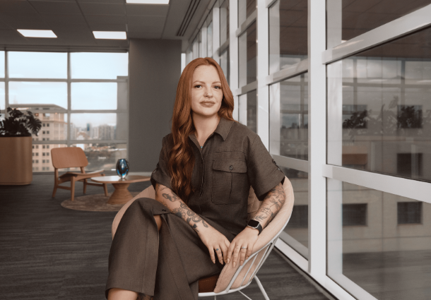A imagem retrata uma mulher de pele clara com longos cabelos ruivos ondulados, sentada em uma cadeira de design moderno em um ambiente de escritório com grandes janelas. Ela veste um macacão marrom de mangas curtas e calças largas, e um smartwatch preto em seu pulso esquerdo. Tatuagens elaboradas são visíveis em seus braços. Ao fundo, através das janelas, observa-se uma vista urbana com prédios e o céu. O escritório parece ter um design contemporâneo com iluminação artificial no teto e luz natural entrando pelas janelas. A atmosfera é profissional e elegante.