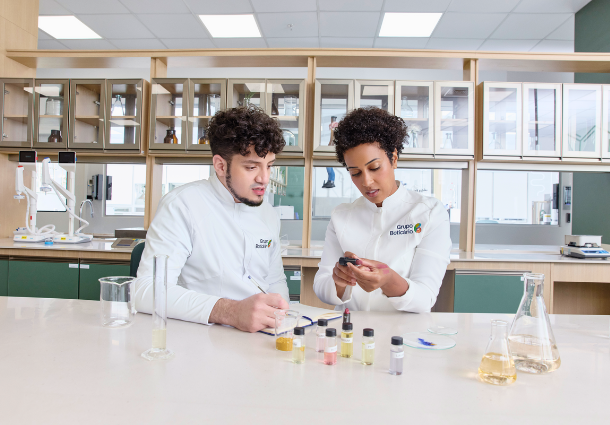 Dois cientistas em jalecos brancos trabalham em um laboratório bem equipado. Um homem escreve em uma prancheta enquanto uma mulher examina algo em suas mãos. À frente deles, há tubos de ensaio com líquidos coloridos e frascos de laboratório sobre uma bancada branca. O ambiente sugere pesquisa e desenvolvimento em um laboratório de inovação, possivelmente na área de alimentos ou ciências biológicas.