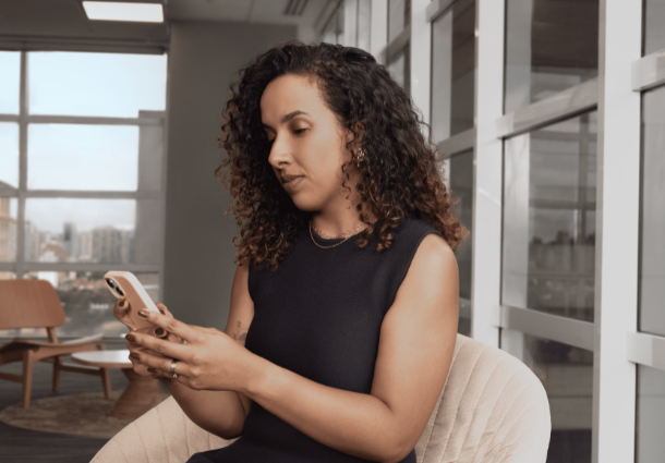 Uma mulher de pele morena clara com cabelos cacheados castanhos está sentada, olhando para o seu smartphone. Ela veste um vestido preto sem mangas e parece concentrada na tela do aparelho que segura com ambas as mãos. Ao fundo, desfocado, percebe-se um ambiente de escritório moderno com janelas amplas e luz natural. Uma cadeira de madeira e uma mesa baixa são visíveis em segundo plano. A cena sugere uma pausa ou um momento de interação digital no ambiente de trabalho.