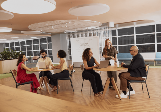 Em um espaço de escritório moderno com piso de madeira clara, dois grupos de pessoas estão reunidos em mesas redondas. O grupo à esquerda, composto por três indivíduos, parece engajado em uma conversa informal com laptops e bebidas sobre a mesa. À direita, um homem careca de óculos e uma mulher ruiva sorridente interagem em outra mesa, também com laptops e bebidas. Ao fundo, uma lousa branca exibe anotações em português, e janelas amplas deixam entrar luz natural. O ambiente sugere uma atmosfera colaborativa e descontraída para discussões de trabalho.