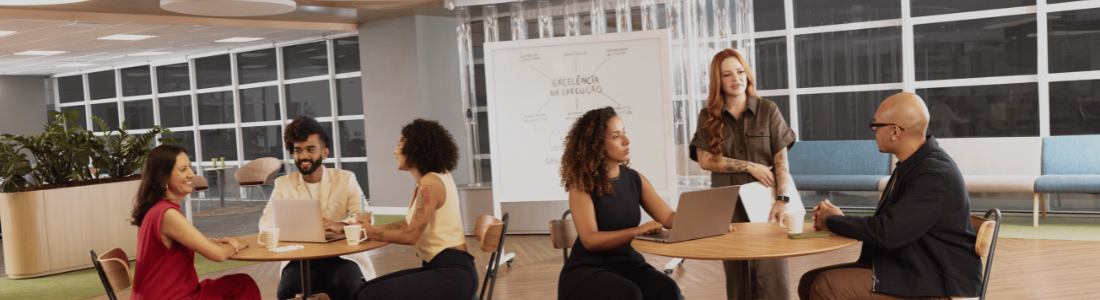 Em um espaço de escritório moderno com piso de madeira clara, dois grupos de pessoas estão reunidos em mesas redondas. O grupo à esquerda, composto por três indivíduos, parece engajado em uma conversa informal com laptops e bebidas sobre a mesa. À direita, um homem careca de óculos e uma mulher ruiva sorridente interagem em outra mesa, também com laptops e bebidas. Ao fundo, uma lousa branca exibe anotações em português, e janelas amplas deixam entrar luz natural. O ambiente sugere uma atmosfera colaborativa e descontraída para discussões de trabalho.