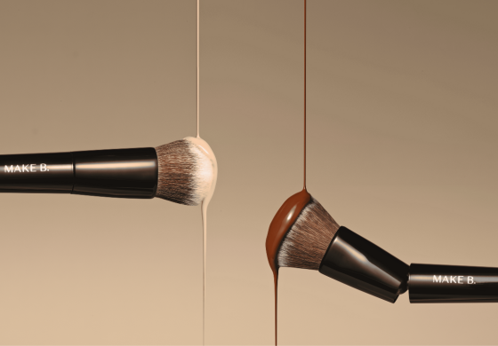 A imagem mostra dois pincéis de maquiagem da marca "Make B." suspensos contra um fundo bege claro. À esquerda, um pincel com cerdas marrom-claras está coberto por uma base líquida bege que escorre de suas cerdas. À direita, outro pincel com cerdas marrom-escuras está coberto por uma base líquida marrom mais escura, que também escorre. Os cabos dos pincéis são pretos e brilhantes, com a inscrição "MAKE B." em letras brancas. A composição sugere uma demonstração da aplicação de diferentes tons de base com os pincéis da marca.