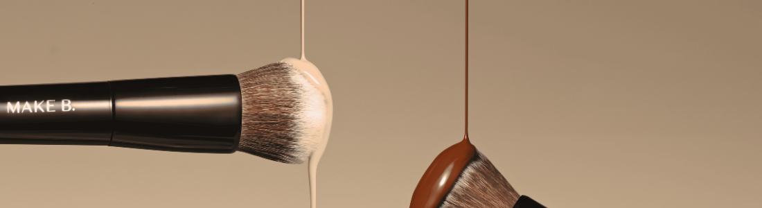 A imagem mostra dois pincéis de maquiagem da marca "Make B." suspensos contra um fundo bege claro. À esquerda, um pincel com cerdas marrom-claras está coberto por uma base líquida bege que escorre de suas cerdas. À direita, outro pincel com cerdas marrom-escuras está coberto por uma base líquida marrom mais escura, que também escorre. Os cabos dos pincéis são pretos e brilhantes, com a inscrição "MAKE B." em letras brancas. A composição sugere uma demonstração da aplicação de diferentes tons de base com os pincéis da marca.