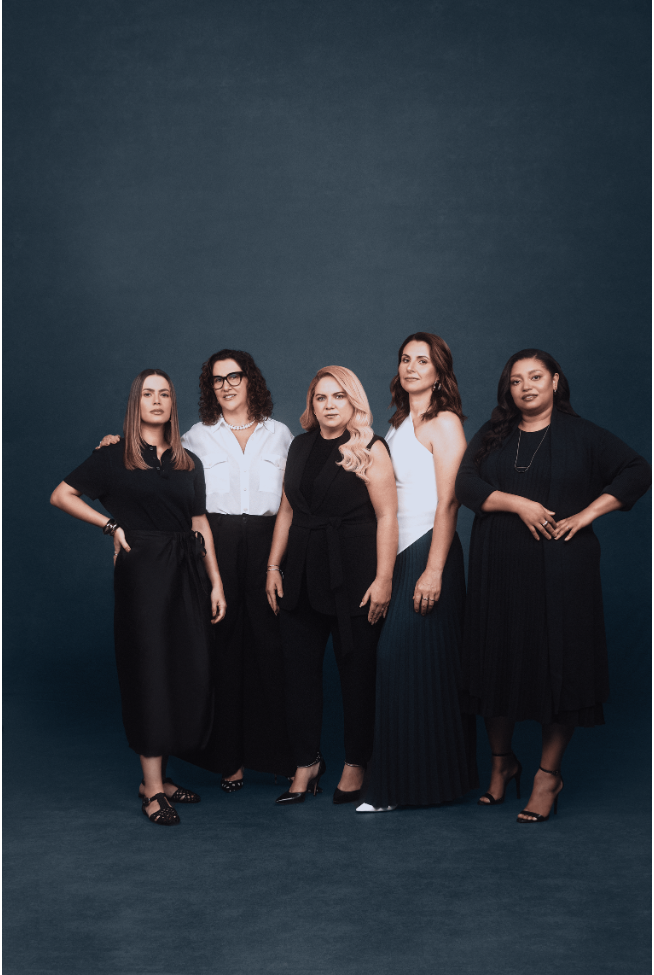 Cinco mulheres diversas posam juntas em frente a um fundo azul-acinzentado escuro. Vestidas predominantemente em tons de preto e branco, elas exibem diferentes estilos e silhuetas. A mulher à esquerda veste um conjunto preto com detalhes drapeados, enquanto a segunda da esquerda usa uma camisa branca clássica e calças pretas. A mulher central opta por um macacão preto elegante, e a penúltima veste um top branco contrastando com uma saia plissada azul escura. À direita, a última mulher veste um vestido preto fluido. A iluminação suave e uniforme foca nas figuras femininas e suas expressões diversas.