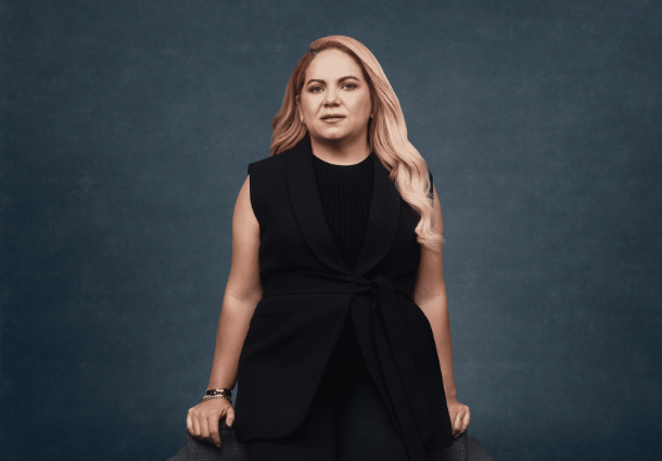 Uma mulher de pele clara com longos cabelos loiros e ondulados está sentada em uma poltrona escura contra um fundo azul-acinzentado. Ela veste um conjunto preto elegante, composto por uma blusa e um colete sem mangas com um detalhe de nó na cintura. Seus braços descansam nos braços da poltrona, e ela usa pulseiras prateadas em ambos os pulsos. Seu olhar é direto e confiante. A iluminação suave e frontal destaca seu rosto e a textura de suas roupas.