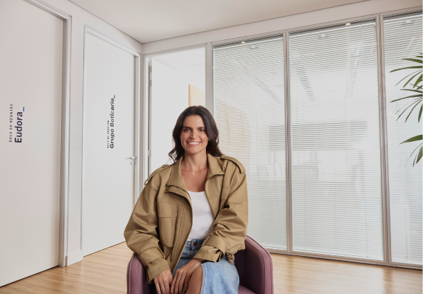 Uma mulher sorridente com cabelos castanhos ondulados está sentada em uma poltrona roxa em um ambiente moderno com piso de madeira clara. Ela veste uma jaqueta bege solta sobre uma camiseta branca e uma saia jeans azul clara. Ao fundo, há grandes janelas com persianas brancas horizontais e uma planta verde alta à direita. Duas portas brancas são visíveis à esquerda, uma com a inscrição vertical "SALA DE REUNIÕES Eudora_" e a outra com "Grupo Boticário_". A luz natural ilumina o espaço, criando uma atmosfera acolhedora e profissional.