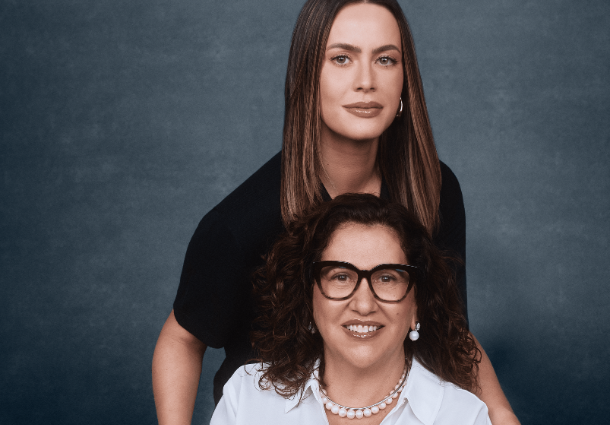 Duas mulheres posam para um retrato em frente a um fundo azul-acinzentado escuro. A mulher em primeiro plano, sentada, tem cabelos castanhos cacheados, usa óculos de armação preta, uma camisa branca de botões e um colar de pérolas. A mulher atrás dela, em pé e ligeiramente inclinada, tem cabelos castanhos lisos e veste uma blusa preta. Ambas sorriem suavemente para a câmera e usam brincos discretos. A iluminação suave e uniforme destaca seus rostos e a proximidade entre elas.