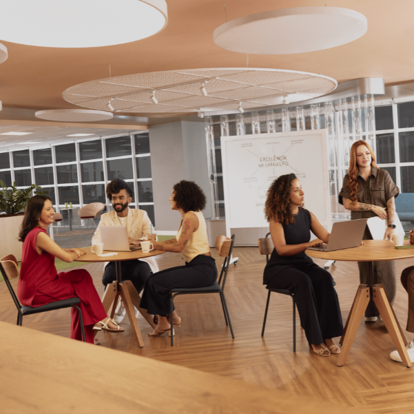 Em um espaço de escritório moderno com piso de madeira clara, dois grupos de pessoas estão reunidos em mesas redondas. O grupo à esquerda, composto por três indivíduos, parece engajado em uma conversa informal com laptops e bebidas sobre a mesa. À direita, um homem careca de óculos e uma mulher ruiva sorridente interagem em outra mesa, também com laptops e bebidas. Ao fundo, uma lousa branca exibe anotações em português, e janelas amplas deixam entrar luz natural. O ambiente sugere uma atmosfera colaborativa e descontraída para discussões de trabalho.