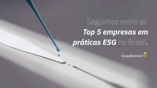 Uma pipeta de vidro inclinada libera uma gota de líquido azul sobre uma superfície branca alongada. A luz cria um forte contraste, projetando sombras nítidas da pipeta e da gota. Ao lado direito, em letras brancas sobre o fundo cinza, lê-se: "Seguimos entre as Top 5 empresas em práticas ESG no Brasil." Abaixo, à direita, aparece o logo "Grupo Boticário". A imagem combina um elemento visual de precisão com uma mensagem corporativa sobre sustentabilidade.