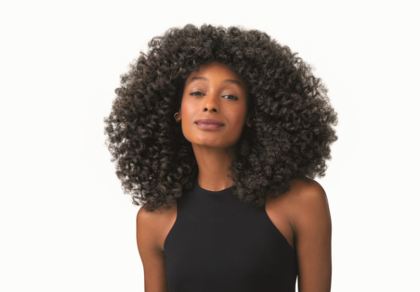 Uma mulher de pele morena clara sorri suavemente para a câmera, emoldurada por um volumoso cabelo afro preto e encaracolado. Ela veste uma blusa preta de gola alta, destacando o contraste com o fundo branco e uniforme. A luz frontal e suave ilumina seu rosto, revelando seus traços delicados e a textura de seus cachos. Seu olhar direto transmite confiança e serenidade. O foco central na mulher e em seu cabelo exuberante cria uma imagem impactante e elegante.