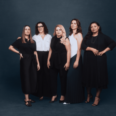 Cinco mulheres diversas posam juntas em frente a um fundo azul-acinzentado escuro. Vestidas em tons predominantemente neutros de preto e branco, cada uma exibe um estilo individual. A mulher à esquerda usa um conjunto preto elegante, enquanto a segunda veste uma camisa branca clássica e calças pretas. No centro, uma mulher loira usa um macacão preto sofisticado. À sua direita, uma mulher veste um top branco com uma saia plissada azul escura, e a última mulher usa um vestido preto fluido. A iluminação suave e uniforme destaca suas expressões confiantes e a variedade de suas personalidades.