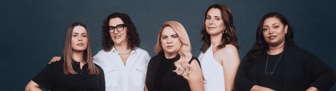 Cinco mulheres diversas posam juntas em frente a um fundo azul-acinzentado escuro. Vestidas em tons predominantemente neutros de preto e branco, cada uma exibe um estilo individual. A mulher à esquerda usa um conjunto preto elegante, enquanto a segunda veste uma camisa branca clássica e calças pretas. No centro, uma mulher loira usa um macacão preto sofisticado. À sua direita, uma mulher veste um top branco com uma saia plissada azul escura, e a última mulher usa um vestido preto fluido. A iluminação suave e uniforme destaca suas expressões confiantes e a variedade de suas personalidades.