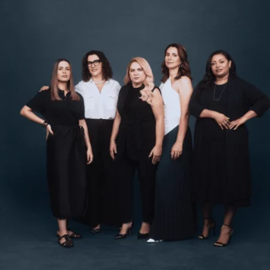Cinco mulheres posam lado a lado em frente a um fundo azul-acinzentado escuro. Vestidas predominantemente em tons de preto e branco, elas exibem diferentes estilos e silhuetas. A mulher à esquerda veste um conjunto preto com detalhes drapeados, enquanto a segunda da esquerda usa uma camisa branca clássica e calças pretas. A mulher central opta por um macacão preto elegante, e a penúltima veste um top branco contrastando com uma saia plissada azul escura. À direita, a última mulher veste um vestido preto fluido. A iluminação suave e uniforme foca nas figuras femininas e suas expressões diversas.
