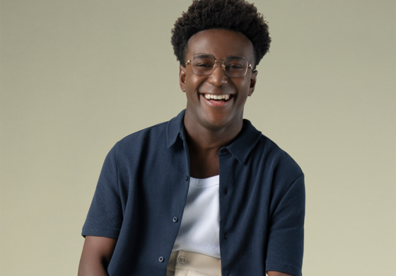 A imagem é um retrato de um jovem negro sorrindo alegremente para a câmera. Ele tem cabelo curto e encaracolado, e usa óculos de armação dourada. Ele veste uma camisa azul escura de manga curta aberta sobre uma camiseta branca. A parte inferior de sua roupa não está totalmente visível, mas sugere uma calça ou bermuda de cor clara. O jovem tem um sorriso largo que mostra seus dentes brancos. Sua expressão facial é de felicidade e entusiasmo. O fundo é um tom bege claro e uniforme, sem distrações, o que foca toda a atenção no sujeito. A iluminação é suave e frontal, realçando suas feições e a vivacidade de seu sorriso.
