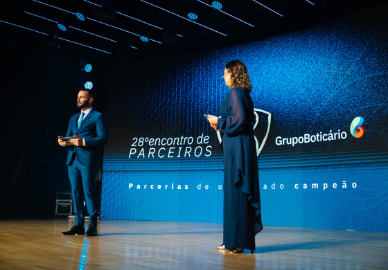 Em um palco iluminado por luzes azuis, um homem de barba e terno azul escuro segura um tablet e sorri para a esquerda. À sua direita, uma mulher de vestido azul escuro e cabelos castanhos encaracolados se dirige para a direita, segurando um microfone. Ao fundo, uma tela grande exibe a inscrição "28º encontro de PARCEIROS" em branco, o logo do Grupo Boticário e um design abstrato em tons de azul. A atmosfera sugere um evento corporativo ou uma apresentação formal. A iluminação destaca os palestrantes contra o cenário digital.