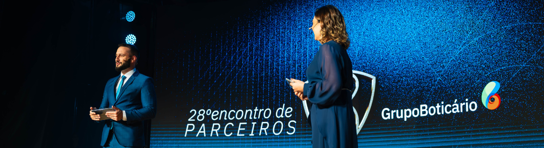 Em um palco iluminado por luzes azuis, um homem de barba e terno azul escuro segura um tablet e sorri para a esquerda. À sua direita, uma mulher de vestido azul escuro e cabelos castanhos encaracolados se dirige para a direita, segurando um microfone. Ao fundo, uma tela grande exibe a inscrição "28º encontro de PARCEIROS" em branco, o logo do Grupo Boticário e um design abstrato em tons de azul. A atmosfera sugere um evento corporativo ou uma apresentação formal. A iluminação destaca os palestrantes contra o cenário digital.