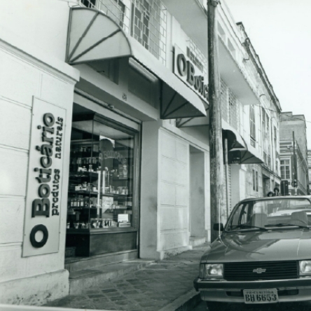Uma fotografia em preto e branco captura a fachada de uma loja O Boticário, identificada por uma placa vertical com a marca e a descrição "produtos naturais". A loja possui uma vitrine onde diversos produtos estão em exposição. Uma marquise listrada em tons claros e escuros se estende sobre a entrada. A loja está localizada em um edifício de dois andares com janelas gradeadas no andar superior. Na rua em frente, um carro está estacionado ao longo da calçada. A arquitetura e o estilo do veículo sugerem que a fotografia foi tirada há algumas décadas.