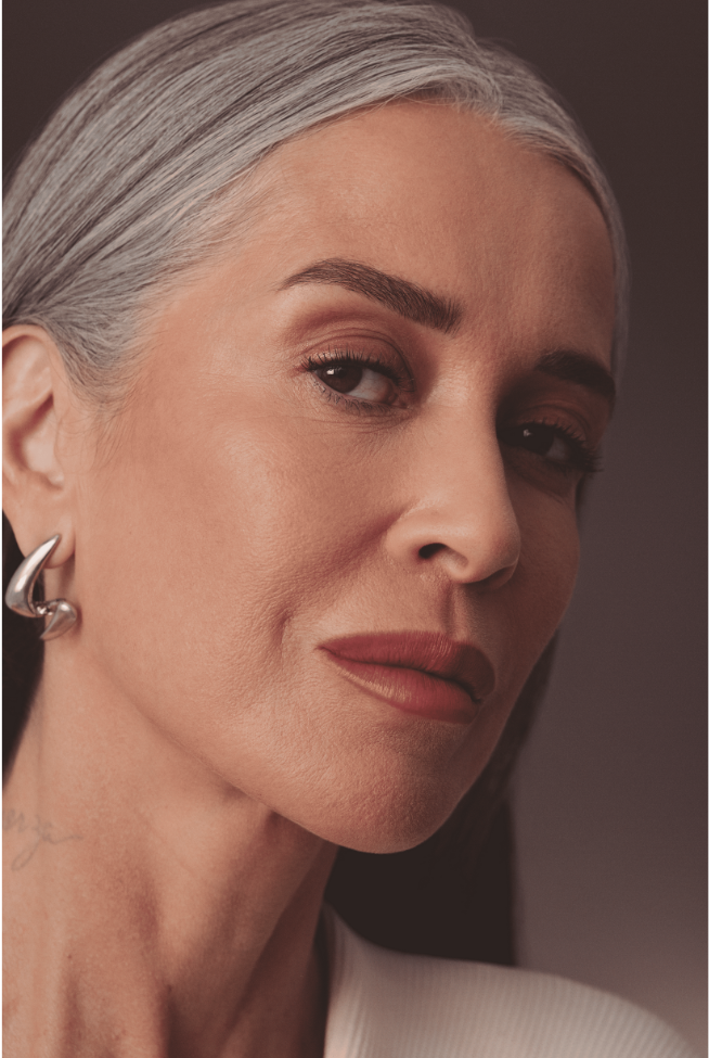 Um close-up do rosto de uma mulher mais velha com cabelos grisalhos e lisos, penteados para trás, revela uma maquiagem sutil. Seus olhos castanhos escuros são levemente realçados, e seus lábios estão pintados com um batom marrom-avermelhado de acabamento levemente brilhante. Ela usa um brinco prateado de design moderno na orelha esquerda. Sua pele mostra sinais de maturidade, com linhas finas visíveis. O fundo é um tom neutro e suave, mantendo o foco em seu rosto expressivo.