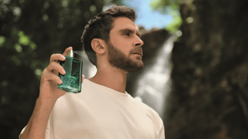 Homem segurando perfume verde ao ar livre, com cachoeira ao fundo e expressão contemplativa.