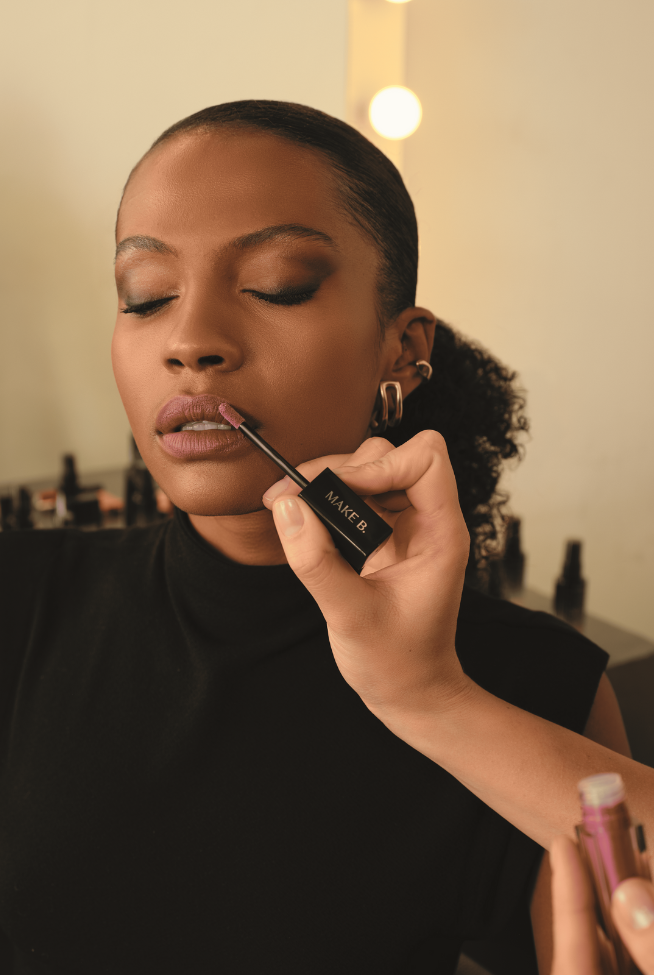 Pessoa aplicando gloss "MAKE B." nos lábios de uma mulher com os olhos fechados.