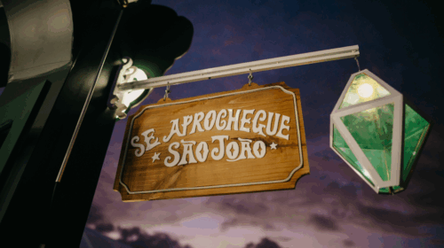 Placa de madeira com os dizeres “Se aprochegue São João” iluminada ao anoitecer.