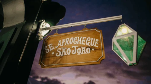 Placa de madeira com os dizeres “Se aprochegue São João” iluminada ao anoitecer.