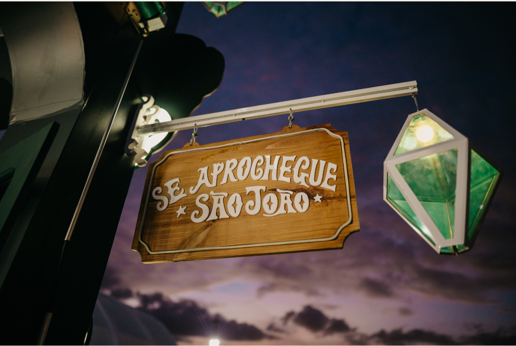 Placa de madeira com os dizeres “Se aprochegue São João” iluminada ao anoitecer.