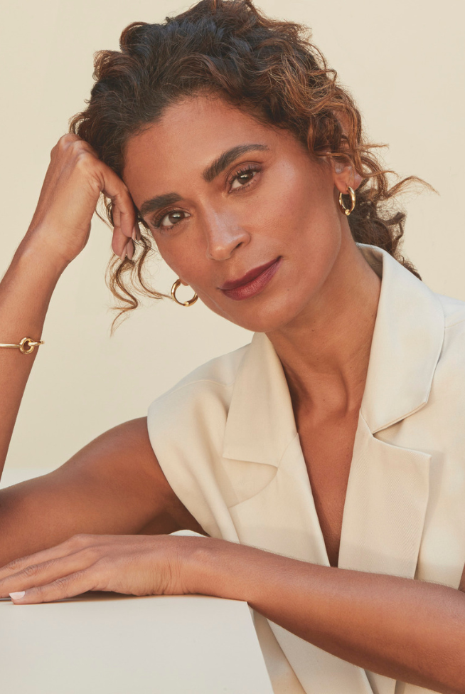 Mulher com expressão serena, usando blazer claro e acessórios dourados, em fundo neutro.
